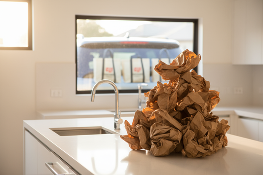Pile de sacs en papier dans une cuisine avec des tote bags oubliés en arrière-plan - illustration humoristique de l'écologie imparfaite